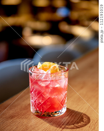 Transparent pink alcohol cocktail with ice, aperol and gin on bar counter in restaurant interior. Alcohol cocktail decorated orange zest spiral. Cold alcoholic cocktail in bar or nightclub. Copy space Transparent pink alcohol cocktail with ice, aperol and gin on bar counter in restaurant interior. Alcohol cocktail decorated orange zest spiral. Cold alcoholic cocktail in bar or nightclub. Copy space 112191019