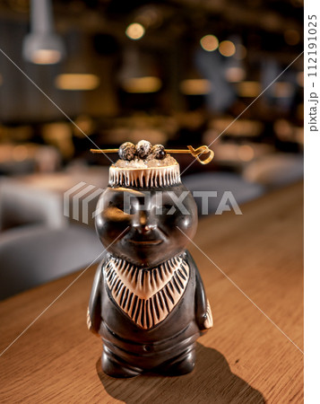 Authentic Tiki Drink Cocktail. Beautiful tropical tiki cocktail on bar counter. Tiki cocktail in monkey mug decorated fig and raspberry on bar in night club or restaurant interior Authentic Tiki Drink Cocktail. Beautiful tropical tiki cocktail on bar counter. Tiki cocktail in monkey mug decorated fig and raspberry on bar in night club or restaurant interior 112191025