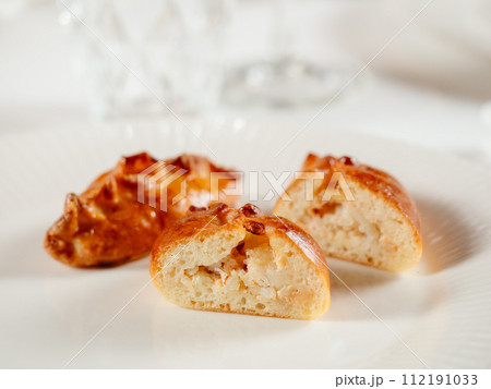 Small russian pies with minced salmon fish Russian Piroshki. Cooked tuna fish hand portioned pies or empanadas cut in half on white plate in elegant restaurant interior. Selective focus, close up. 112191033