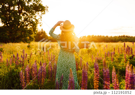 A young woman in a stylish green dress and hat enjoys the sunset in a field with lupins. 112191052