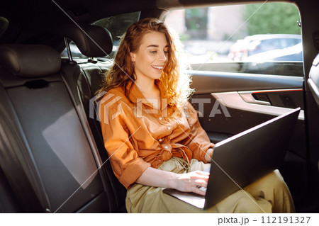 A young curly woman with a laptop sitting in back seat of a car. Beautiful woman uses laptop in car. 112191327