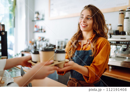 Takeaway food concept. A beautiful female barista gives to-go coffee to a client. Business concept. 112191408