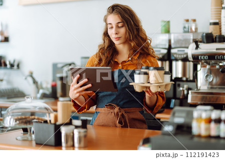 Handsome manager of coffee shop with curly hair tablet in hands gives takeaway coffee in coffee shop Handsome manager of coffee shop with curly hair tablet in hands gives takeaway coffee in coffee shop 112191423