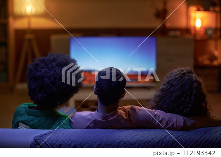 Back view of three people sitting on couch together and watching TV in dark room Back view of three people sitting on couch together and watching TV in dark room 112193342