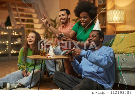 Full length portrait of multiethnic group of adults playing video games emotionally during party at home with African American woman holding controller Full length portrait of multiethnic group of adults playing video games emotionally during party at home with African American woman holding controller 112193385