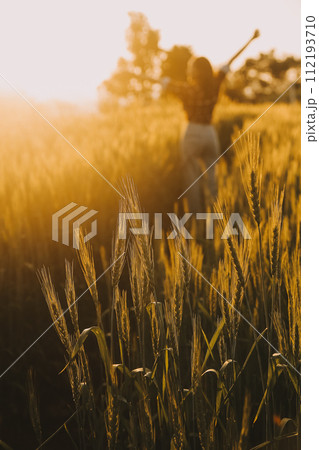 Young pretty woman in red summer dress and straw hat walking on yellow farm field with ripe golden wheat enjoying warm evening. 112193710