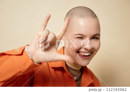 Portrait of carefree bald young woman with rock hand sign smiling at camera in studio and having fun 112193862