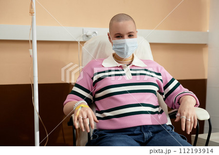 Front view portrait of bald young woman wearing mask getting IV drip treatment in clinic and smiling at camera copy space 112193914
