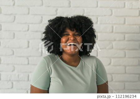 Laughing African American woman with an afro hairstyle and good sense of humor smiling and laugh on brick wall at home background. Happiness and good emotions concept 112196234