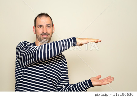 Bearded Hispanic man wearing a striped sweater with his arms open in profile offering something while smiling at the camera, isolated on beige background Bearded Hispanic man wearing a striped sweater with his arms open in profile offering something while smiling at the camera, isolated on beige background 112196630