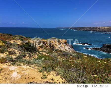 A coastal landscape with rocky cliffs, greenery, and a clear blue sky. The ocean waves crash against the rocks. 112196888
