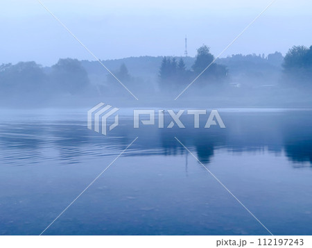 Morning fog on the lake in the early misty morning. The trees are reflected in the water. 112197243