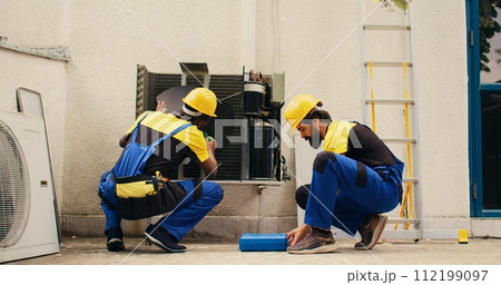 Teamworking competent mechanic opening up rusty out of service HVAC system to replace it with new performant outside condenser after draining refrigerant and replacing ductwork 112199097