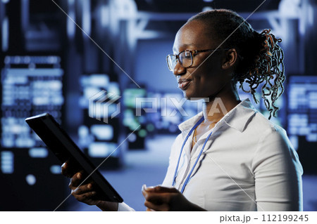 Upbeat admin in data center uses tablet to prevent system infrastructure overload. Precise expert between blade servers ensuring enough network bandwidth for smooth operations 112199245