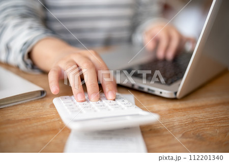 A close-up image of a woman using a calculator while working on her laptop computer at a table. 112201340