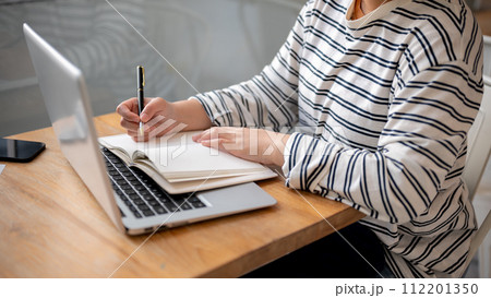 A woman working remotely at a cafe, taking notes in her notebook while working on her laptop. A woman working remotely at a cafe, taking notes in her notebook while working on her laptop. 112201350