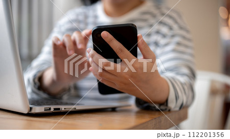 A woman using her smartphone, responding to messages, while working remotely at a cafe. 112201356