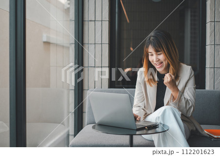 Joyful young businesswoman with laptop sitting in a modern office space, celebrating a successful moment. 112201623