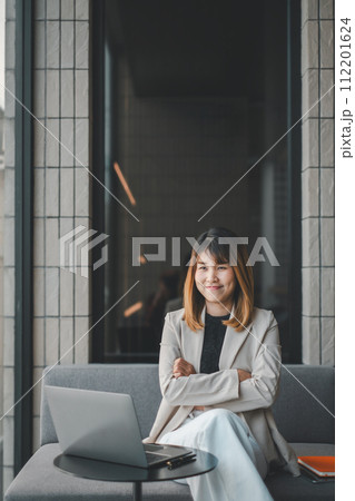 A content young Asian businesswoman sits on a couch in a modern office setting, arms crossed, with her laptop and smartphone nearby, reflecting a successful professional moment. 112201624