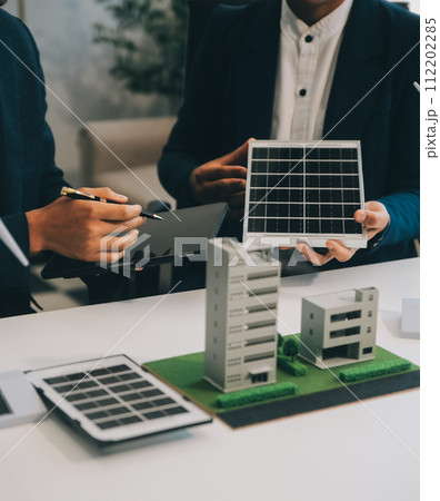 Two Young Engineers Expertise in Solar Cell Installation Meetings and Discussion in The Job. Planning to Install Solar Photovoltaic Panels on Roof Top in The Office Room with Factory Building Plan. 112202285