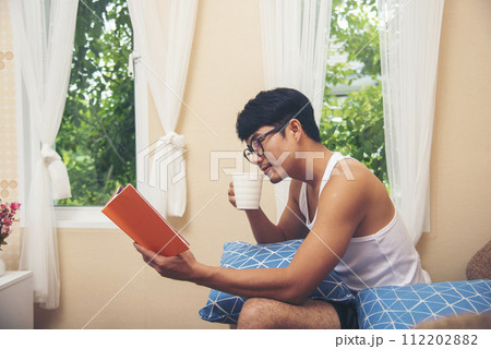 Young asian man reading book on cozy couch sofa at warm home. Relax man holding book read on sofa happiness. Man reading open book leisure mind in living room. Happy enjoyment man lifestyle at home 112202882