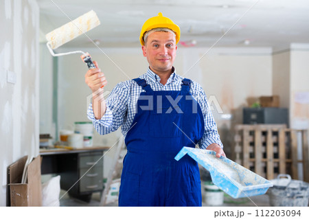 Portrait of positive man painter in a protective helmet and overal with paint roller Portrait of positive man painter in a protective helmet and overal with paint roller 112203094