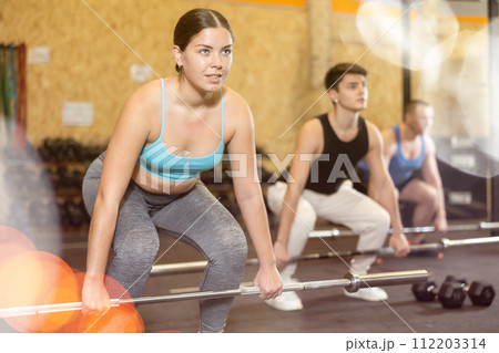 Young dedicated woman doing sit-ups with rod among other people in gym Young dedicated woman doing sit-ups with rod among other people in gym 112203314