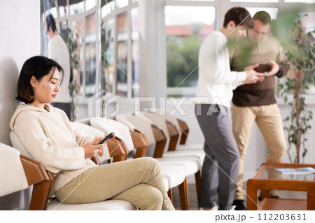 Girl sits in lobby of employment office, waiting for interview or conversation with career consultant. Watches news on Internet via smartphone 112203631