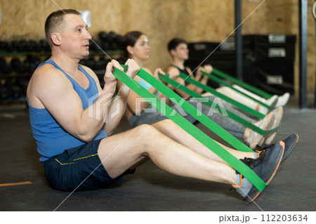Motivated man practicing exercises with stretch rope sitting near other people in gym 112203634