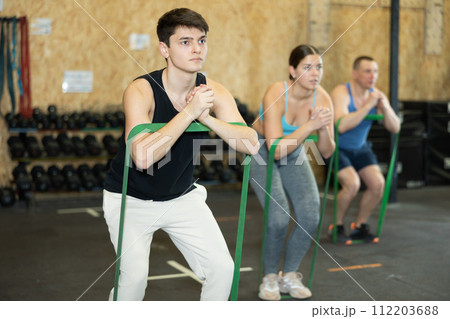 Motivated young man practicing exercises with stretch rope standing near other people in gym 112203688