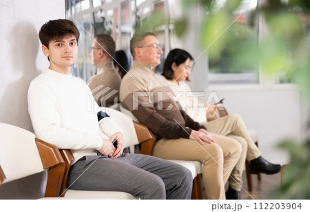Young man browsing websites on smartphone while sitting in lobby of job center or employment office, waiting for interview or conversation with career counselor Young man browsing websites on smartphone while sitting in lobby of job center or employment office, waiting for interview or conversation with career counselor 112203904