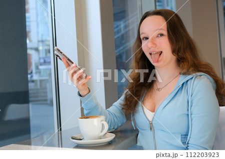 stick out tongue young teenage girl sits by the window with coffee and phone in hands looks into the frame showing tongue funny photos interesting emotion adolescence young woman 112203923