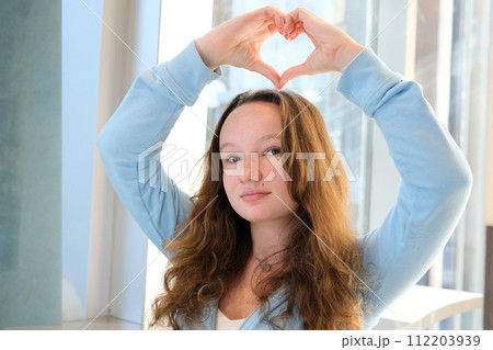 tender european girl makes heart with hands smiles sits at table by the window smile joy happiness convey love show heart on right at left and at top space for text advertising blue sports blouse 112203939