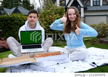 lovely picnic boys teenage girls holding laptop with green screen chromakey advertising boxes of pizza girl showing two thumbs up sitting on plaid private sector relaxing learning online outdoors 112203970