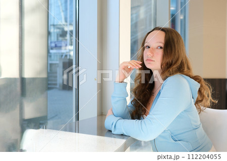Depressed, sad woman at home looking out window for mental health, depression or heartbreak. Young gen z girl alone in apartment with lonely or bored face and thinking of problem or future life 112204055
