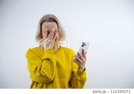 Surprised pretty European woman with blond hair covering her mouth with her hand and looking at her phone, receiving a text, posing against a soft purple background. Human emotions, reaction concept Surprised pretty European woman with blond hair covering her mouth with her hand and looking at her phone, receiving a text, posing against a soft purple background. Human emotions, reaction concept 112204271