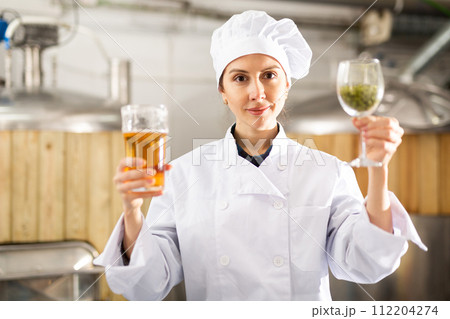Woman brewer holding glass of hops and glass of beer in her hands at brewery Woman brewer holding glass of hops and glass of beer in her hands at brewery 112204274