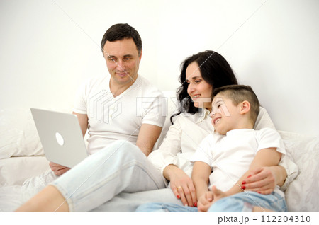 bed linen advertising family watching movie on laptop on bed relaxing together white clothes chatting online with relatives mom dad and son laughing smiling rejoicing happy life 112204310