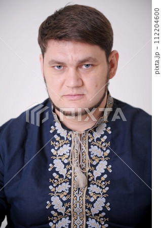 stern look of blue eyes handsome man in Ukrainian embroidered shirt looks straight into frame he has light beard compressed lips thick hair a blue shirt and embroidery on a white background close-up 112204600