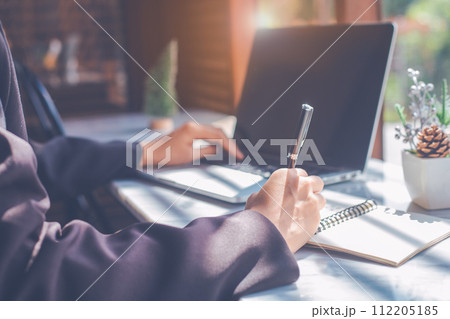 Business woman hand writing on a notepad with a pen and use a laptop computer in the office. 112205185