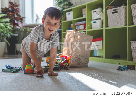 A cheerful European boy plays with cars on the carpet in his room. The child dumped the toys 112207987
