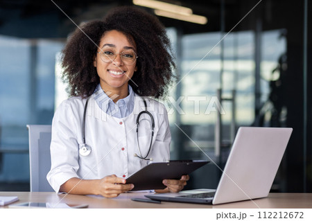 Portrait of a smiling African-American female doctor sitting in a white coat at the desk in the office, working on a laptop, holding a folder with patient case histories, looking into the camera. 112212672