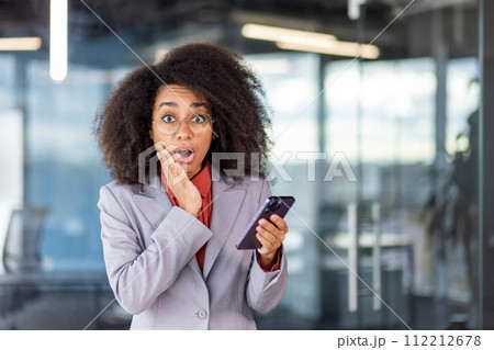 Portrait of shocked young African American woman in business suit standing in office, holding mobile phone, looking at camera in surprise, holding hand to face. Portrait of shocked young African American woman in business suit standing in office, holding mobile phone, looking at camera in surprise, holding hand to face. 112212678