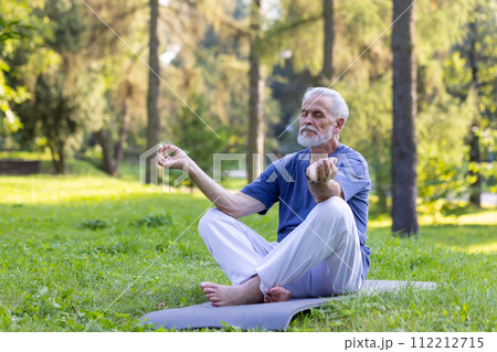 Senior gray-haired man doing yoga in the park, sitting on a mat in the lotus position and meditating with closed eyes alone. 112212715