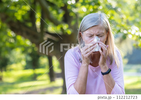 Close-up photo of a senior gray-haired woman standing outside in a park and wiping her nose with a napkin, suffering from a runny nose and seasonal allergies. Close-up photo of a senior gray-haired woman standing outside in a park and wiping her nose with a napkin, suffering from a runny nose and seasonal allergies. 112212723