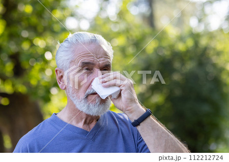 Close-up portrait of a sick elderly man with a beard standing in a park in nature, wiping his nose with a napkin, suffering from a runny nose and seasonal allergies. 112212724