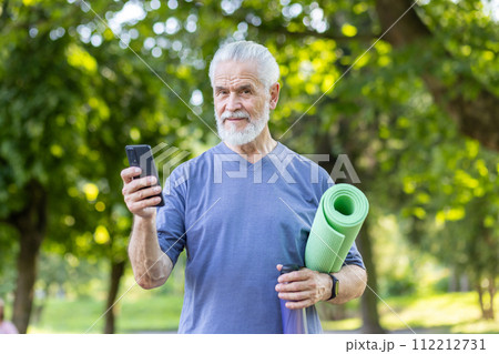 Portrait of a smiling and healthy gray-haired man standing in the park, holding a phone, a yoga mat and a bottle of water, confidently looking at the camera. 112212731