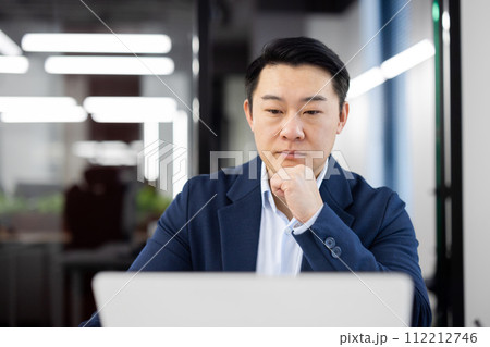 Close-up photo of a focused and serious young Asian man working in the office at a laptop and looking thoughtfully at the screen. 112212746