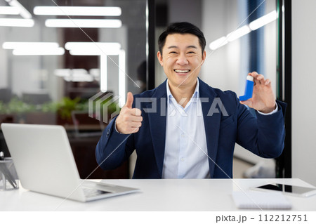 Portrait of a smiling young Asian man in a business suit sitting in the office, holding an asthma and allergy inhaler in his hand, and showing the camera a super gesture with his finger. 112212751