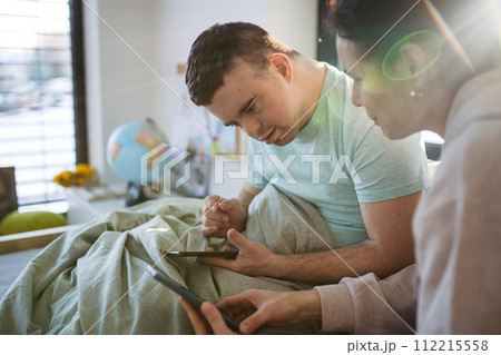 Young man with down syndrome sitting in bed with mom, looking at smartphone in morning. Morning routine for man with disability. 112215558